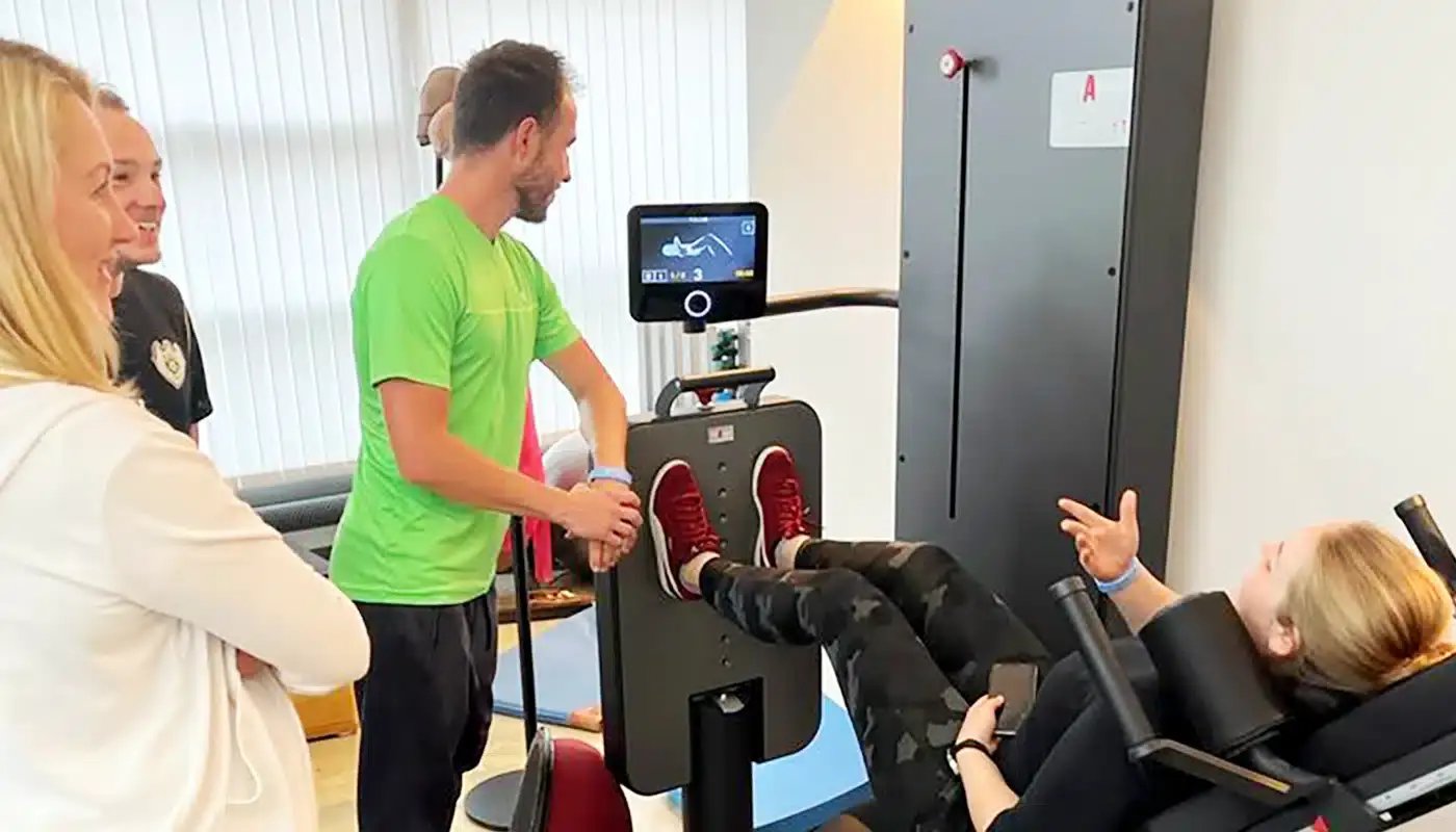 Physiotherapeut demonstriert Übung an einem Trainingsgerät mit liegender Patientin